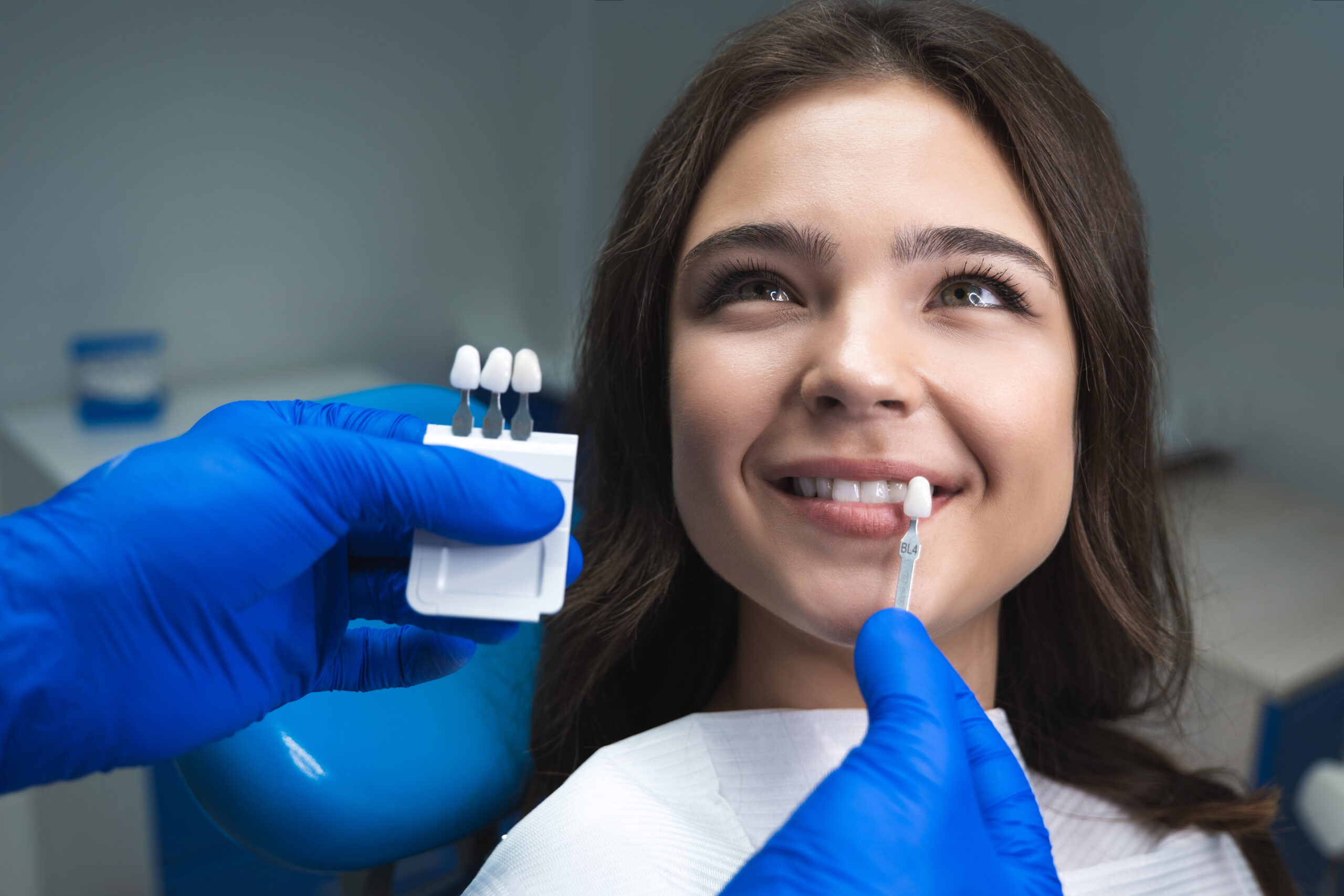 dentist holding porcelain veneers up against young woman's natural teeth