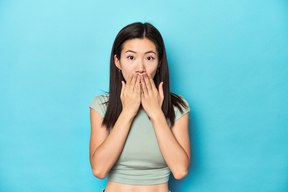 Asian woman in summer green top, studio backdrop, shocked, covering mouth with hands, anxious to discover something new.