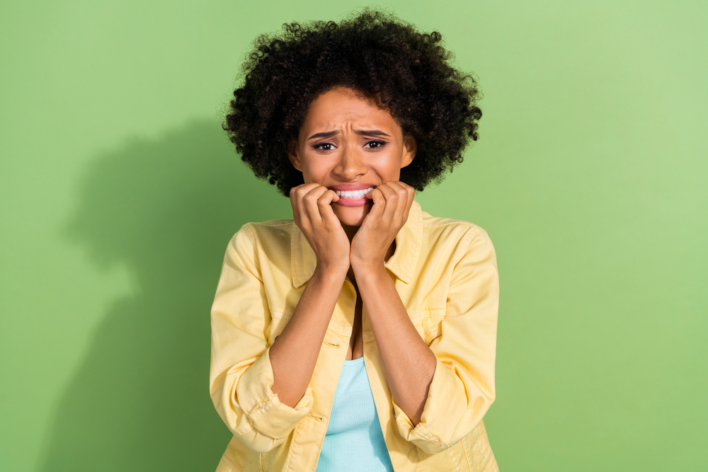 woman holding her face and jaw because of jaw pain, wearing a yellow shirt, on green background