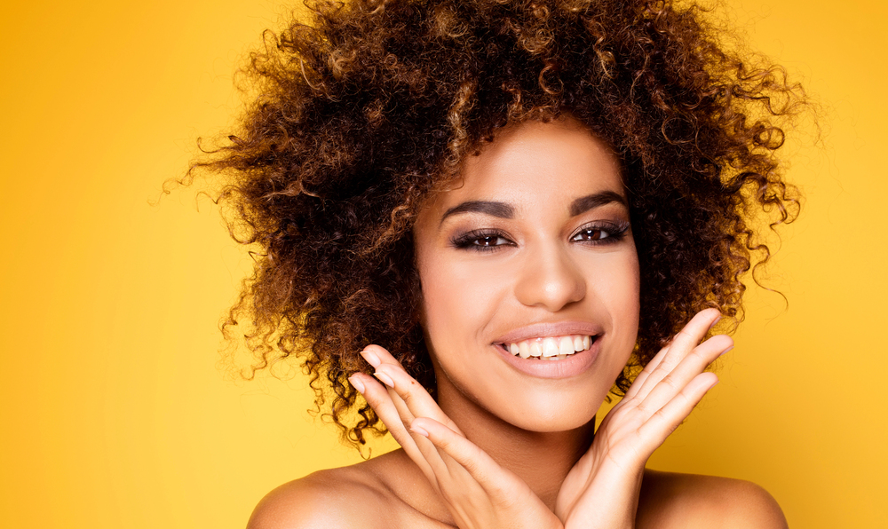 beautiful black woman holding hands in a V-shape under her chin showing perfect smile on yellow background