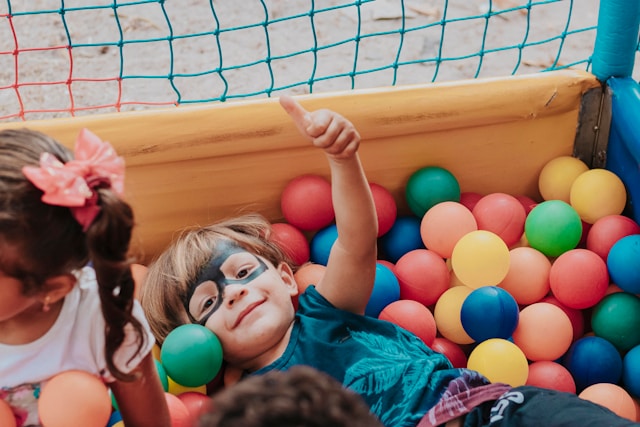 young boy wearing a super hero mask, laying in a ball pit, holding up his thumb and smiling