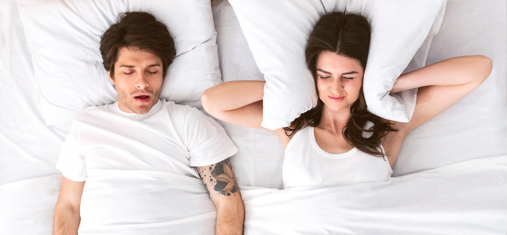 a couple laying in bed, woman covering her ears with a pillow, man sleeping with mouth open and snoring because of sleep apnea